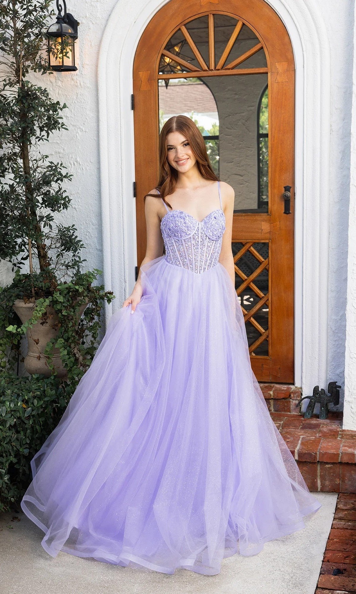 A young woman stands before a wooden door wearing the Portia and Scarlett XO163 prom ball gown in lilac, featuring a long silhouette, fitted embellished bodice, and thin straps.